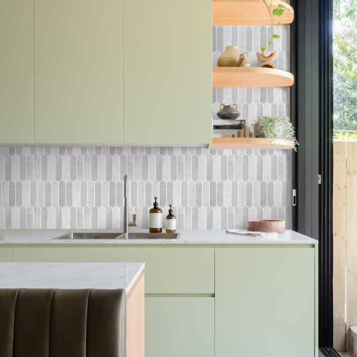 A modern kitchen interior with pale sage green cabinets, a white marble countertop, a gray and white geometric mosaic tile backsplash, open wood floating shelves with decorative items, and a large black-framed glass door on the right.