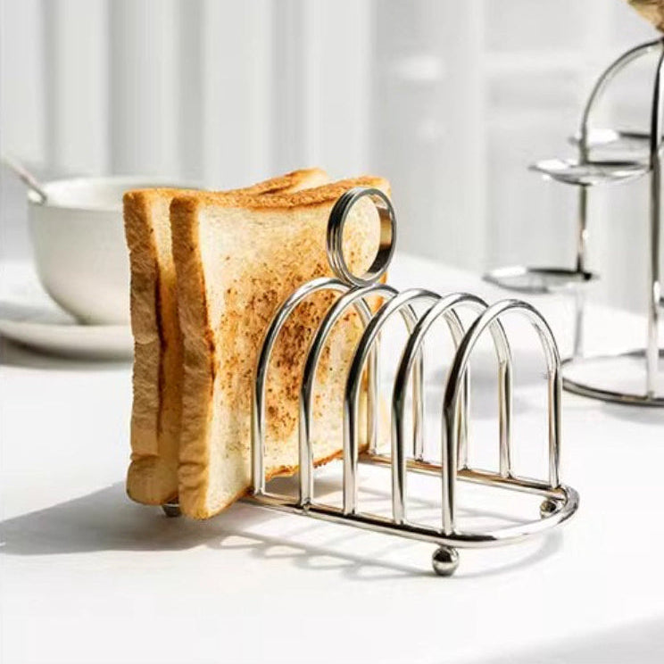 A stainless steel 5-slice toast rack holding two slices of toasted bread, set on a white table with a softly blurred kitchen background.