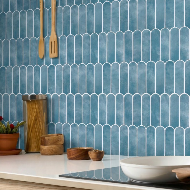 Blue arched ceramic wall tiles with white grout lines on a kitchen backsplash, styled with wooden utensils, a pasta jar, bowls, and a white stovetop pan.