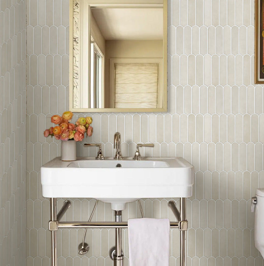 A bathroom vanity setup featuring a white rectangular pedestal sink with a chrome metal stand, a gold-framed mirror on a patterned beige wall, and a vase of orange flowers.