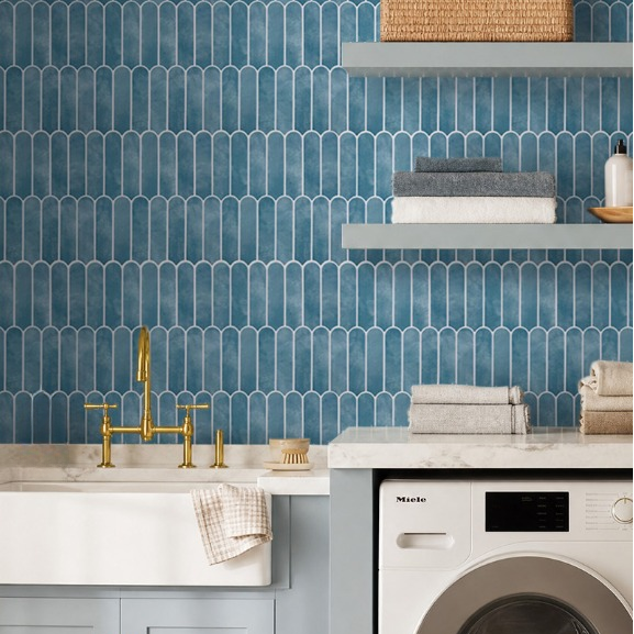 A styled laundry room corner featuring a blue hexagonal tile backsplash, a white farmhouse sink with a gold faucet, light blue cabinets, a white Miele washing machine, and floating shelves with towels and a woven basket.