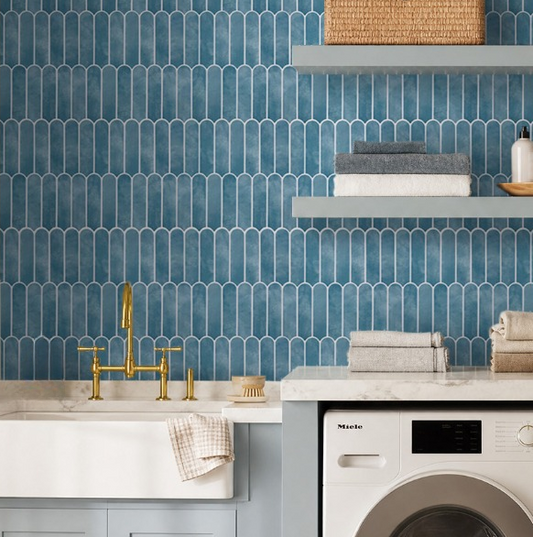 A styled laundry room corner featuring a blue hexagonal tile backsplash, a white farmhouse sink with a gold faucet, light blue cabinets, a white Miele washing machine, and floating shelves with towels and a woven basket.