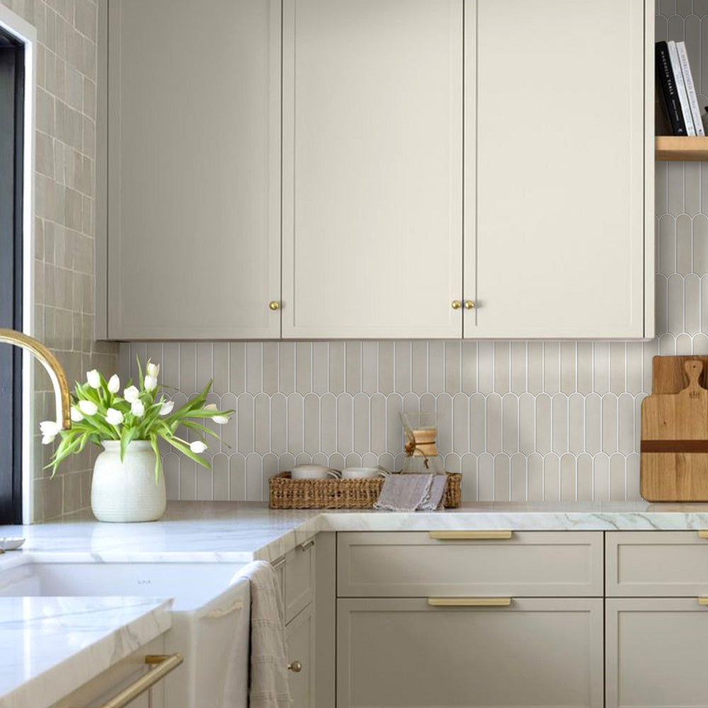 A modern kitchen interior with light beige cabinets, a white marble countertop, a beige subway tile backsplash, and brass hardware. A white vase with tulips sits on the counter next to a window.