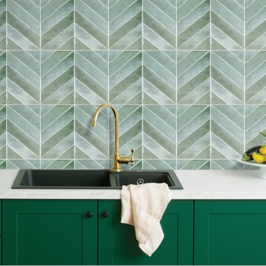 A kitchen backsplash made of light green and white chevron-patterned ceramic tiles, installed above a white countertop with a black double sink, a gold faucet, and dark green cabinets.